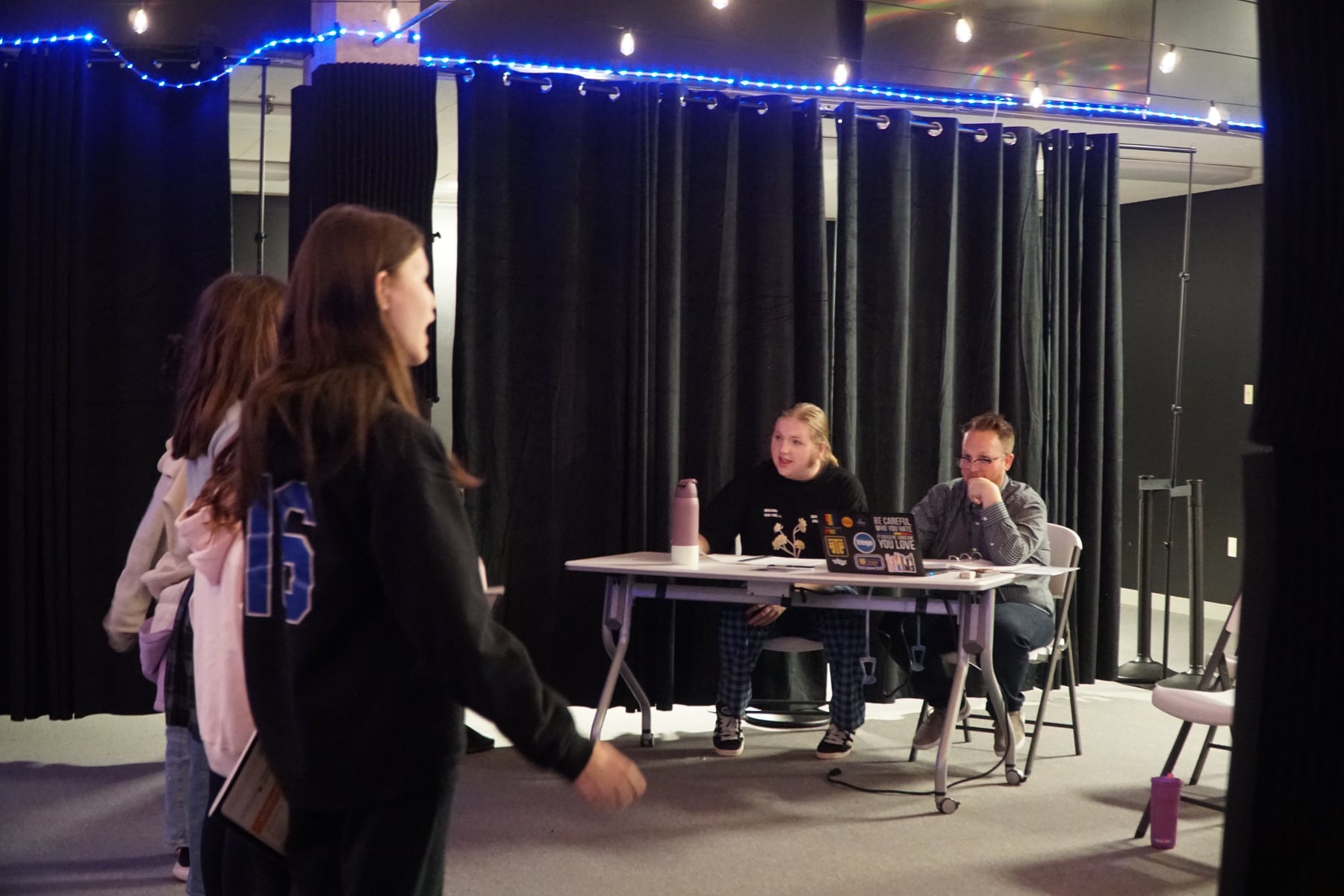 A school musical audition in progress: a student mid-slate on the mark, sheet music in hand, with the director and an accompanist listening from behind the audition table