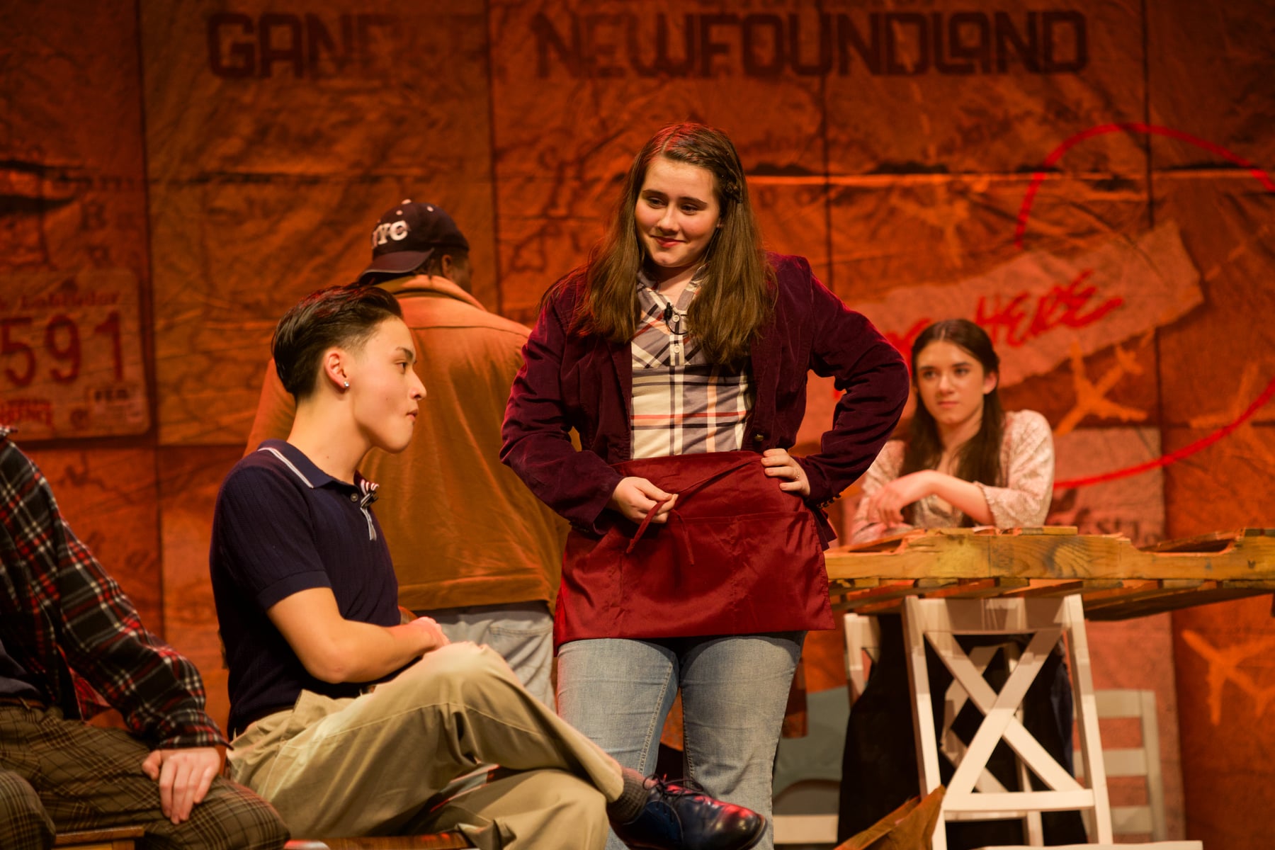 Four high school students in a scene onstage, one in an apron with her hands on her hips, in a school production of Come From Away