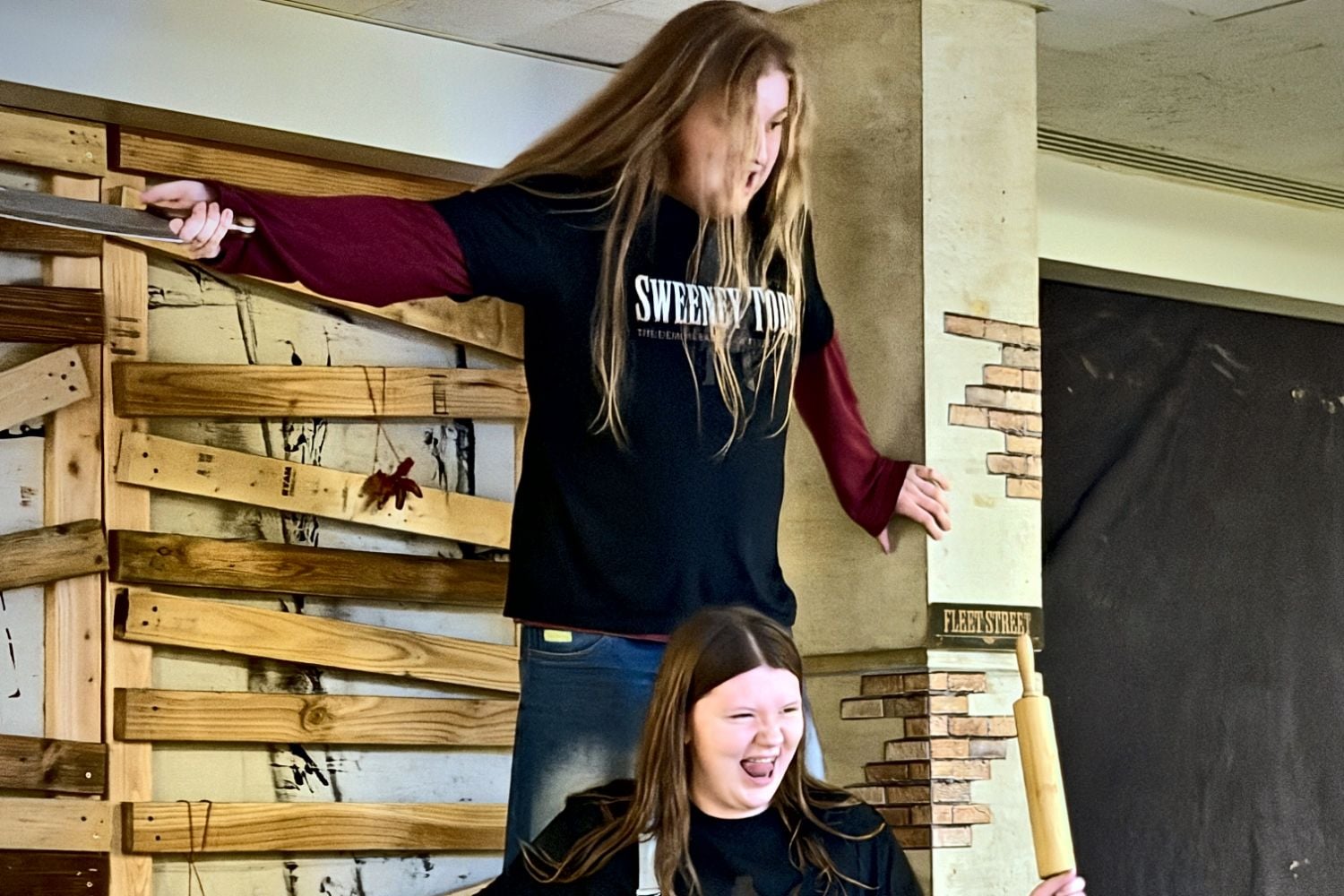 Two Teen Conservatory students in Sweeney Todd shirts laughing on the Fleet Street set during rehearsal