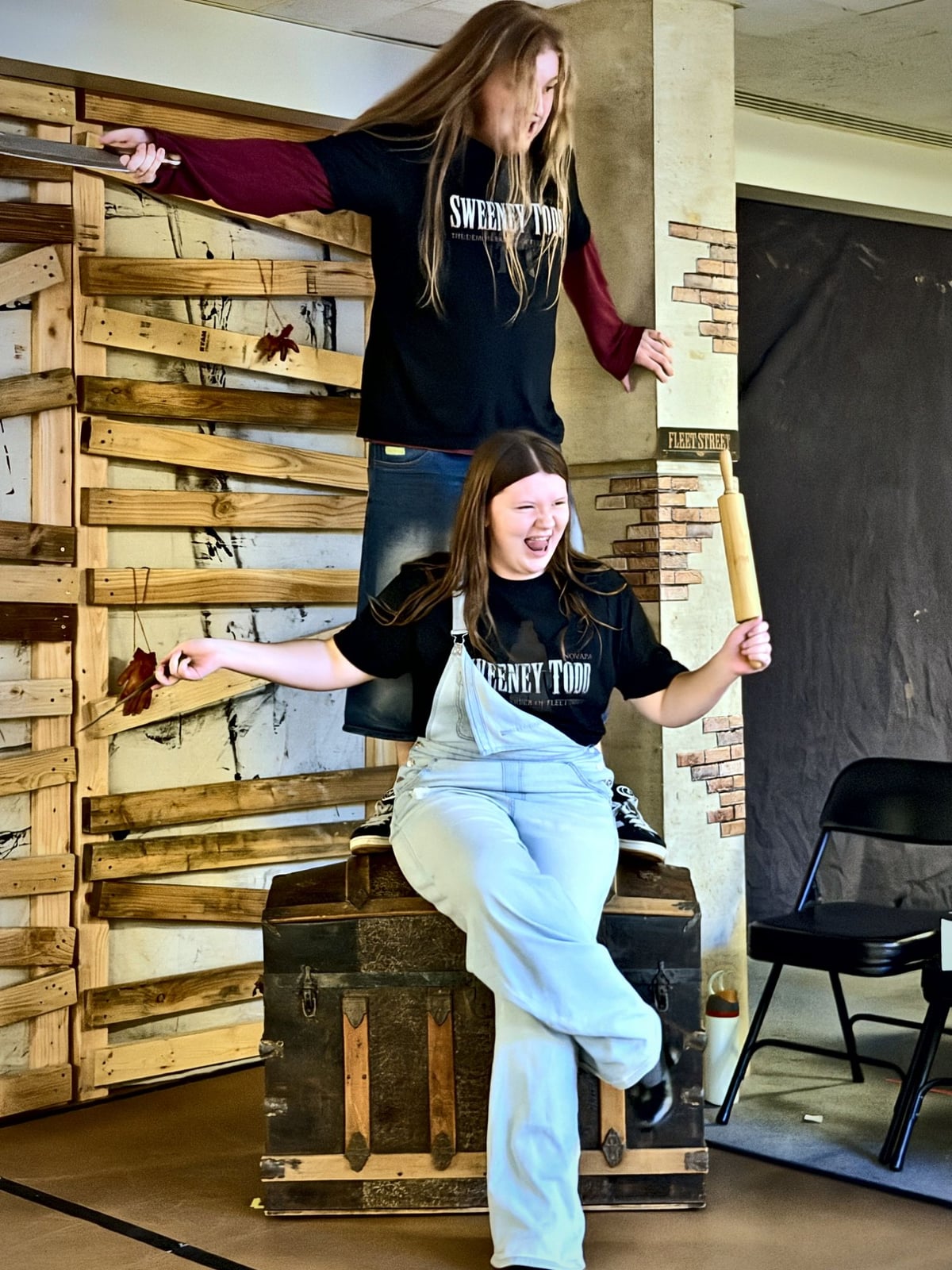 Two Teen Conservatory students in Sweeney Todd shirts clowning on the Fleet Street set during rehearsal, one standing with a prop razor and one seated holding a rolling pin, both laughing